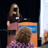 Shot of scholar wearing mask giving oral presentation from behind a few audience members.